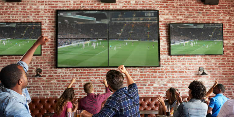 Photo of people at a restaurant cheering while watching large TV screens showing a soccer game.