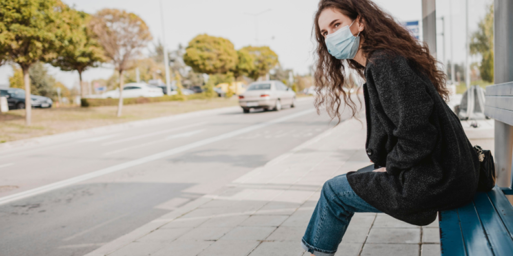 A woman wearing a safety mask waiting at a bus stop