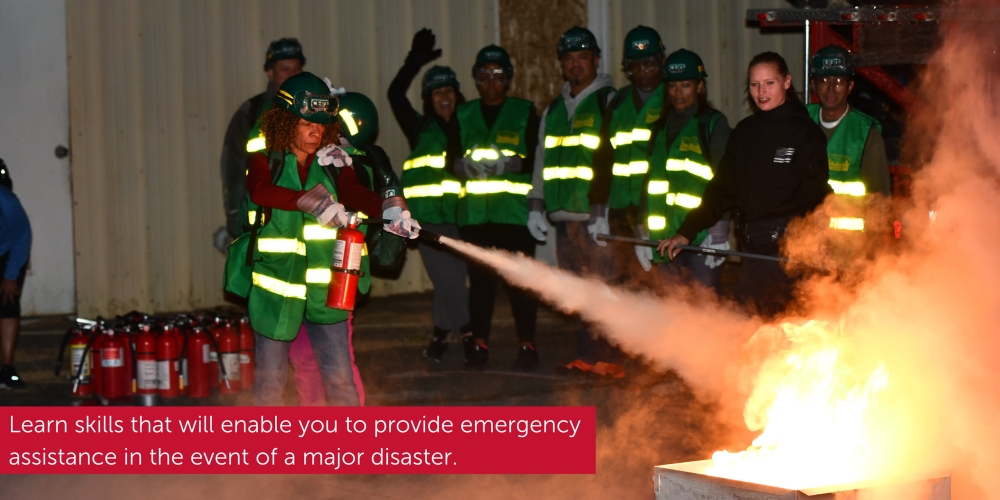 A woman wearing green CERT safety gear uses a fire extinguisher to put out flames