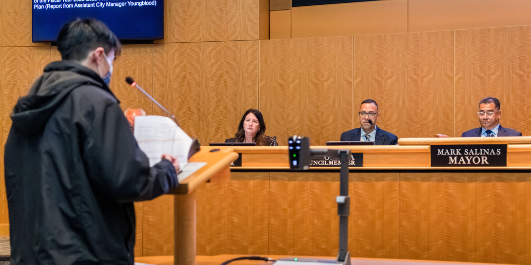 A person wearing a face mask standing at the podium in council chambers addressing the City Council